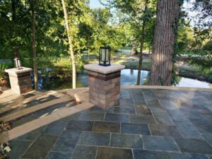 image000001 - October Hardscaping Stone-tiled patio with brick pedestals and lanterns, overlooking a pond framed by trees in a park.