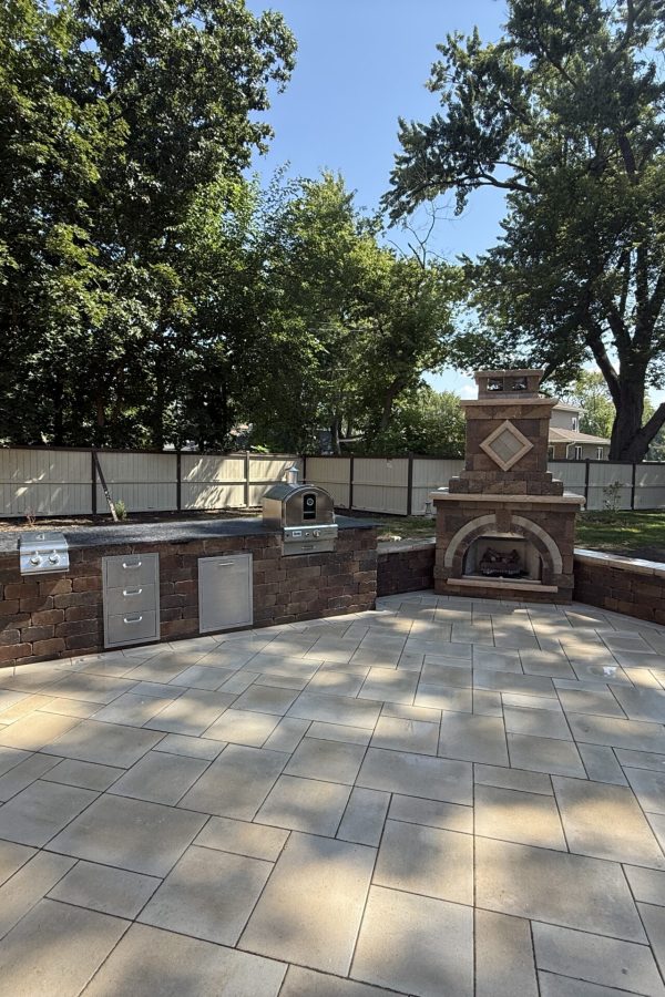 Outdoor kitchen with a brick grill, stainless steel appliances, and a brick oven on a tiled patio; a wooden fence and trees in the background.